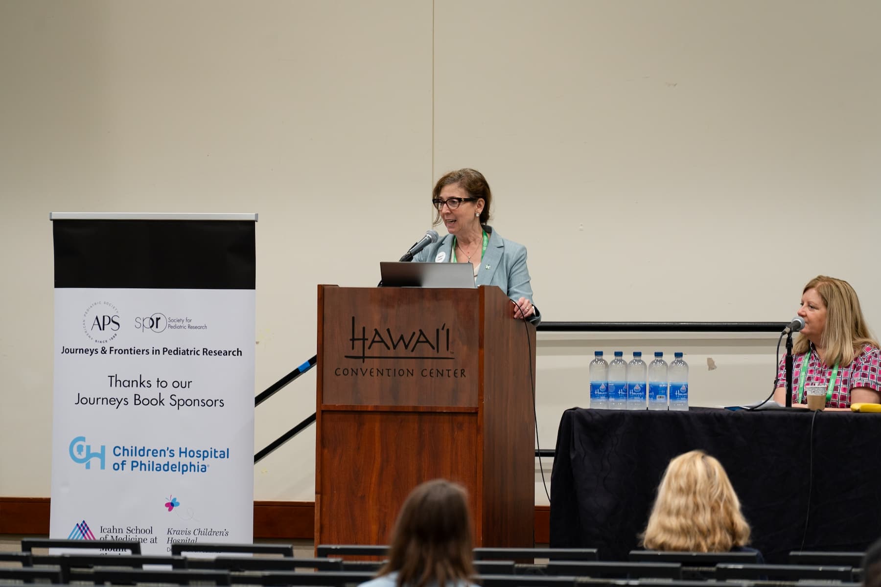 A speaker standing at a wooden podium labeled “Hawai‘i Convention Center,” delivering a presentation in a conference room. A banner to the left displays logos and text thanking sponsors, including the Children’s Hospital of Philadelphia. On the right, another person is seated at a table with several water bottles. Rows of chairs and attendees are visible in the foreground.
