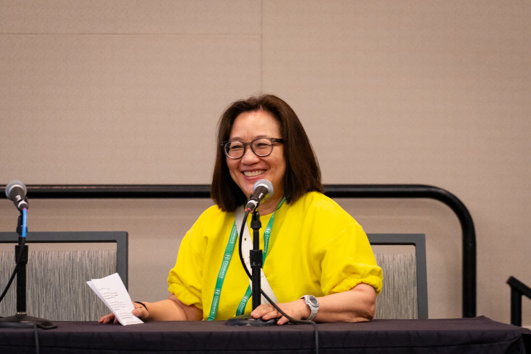 A person seated at a conference table speaking into a microphone, holding a sheet of paper. The individual is wearing a bright yellow top and a green lanyard. Two microphones and water bottles are placed on the table, and the background features a plain beige wall.