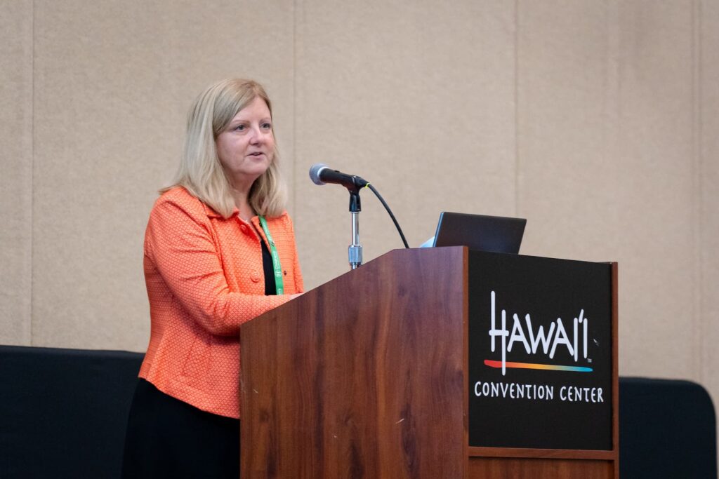 Individual standing at a podium labeled “Hawai‘i Convention Center,” delivering a presentation. The person is wearing an orange textured jacket over a black outfit, with a green lanyard visible. A microphone and a laptop are positioned on the podium, and the background is a plain beige wall.