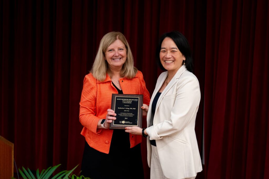 Two individuals standing indoors in front of a deep red curtain. One person is wearing an orange textured jacket over a black top and holding a rectangular award plaque with gold text. The other person is dressed in a light-colored suit jacket over a dark top and is also holding the plaque. The setting appears formal, likely an award presentation or recognition event.