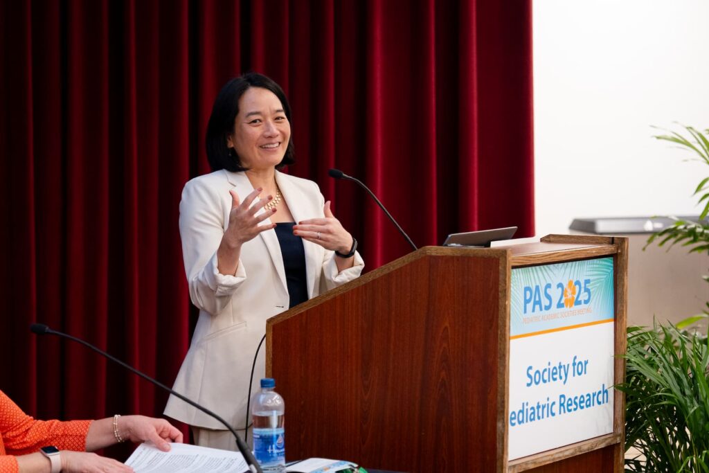 Speaker presenting at a podium during PAS 2025 conference, wearing a light-colored blazer and dark top, with a microphone and water bottle visible; the podium displays the text “PAS 2025 Society for Pediatric Research” and a red curtain is in the background.