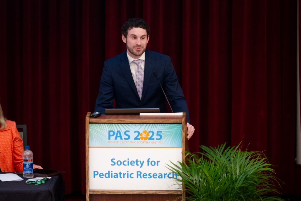 Person standing at a podium with a sign reading “PAS 2025 Society for Pediatric Research” in front of a red curtain, with a water bottle and papers on a nearby table.