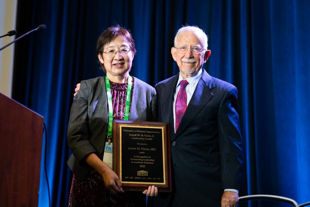 Two individuals standing together in front of a blue curtain backdrop at a conference; one person is holding a large plaque with gold text, and both are wearing formal attire with visible lanyards.