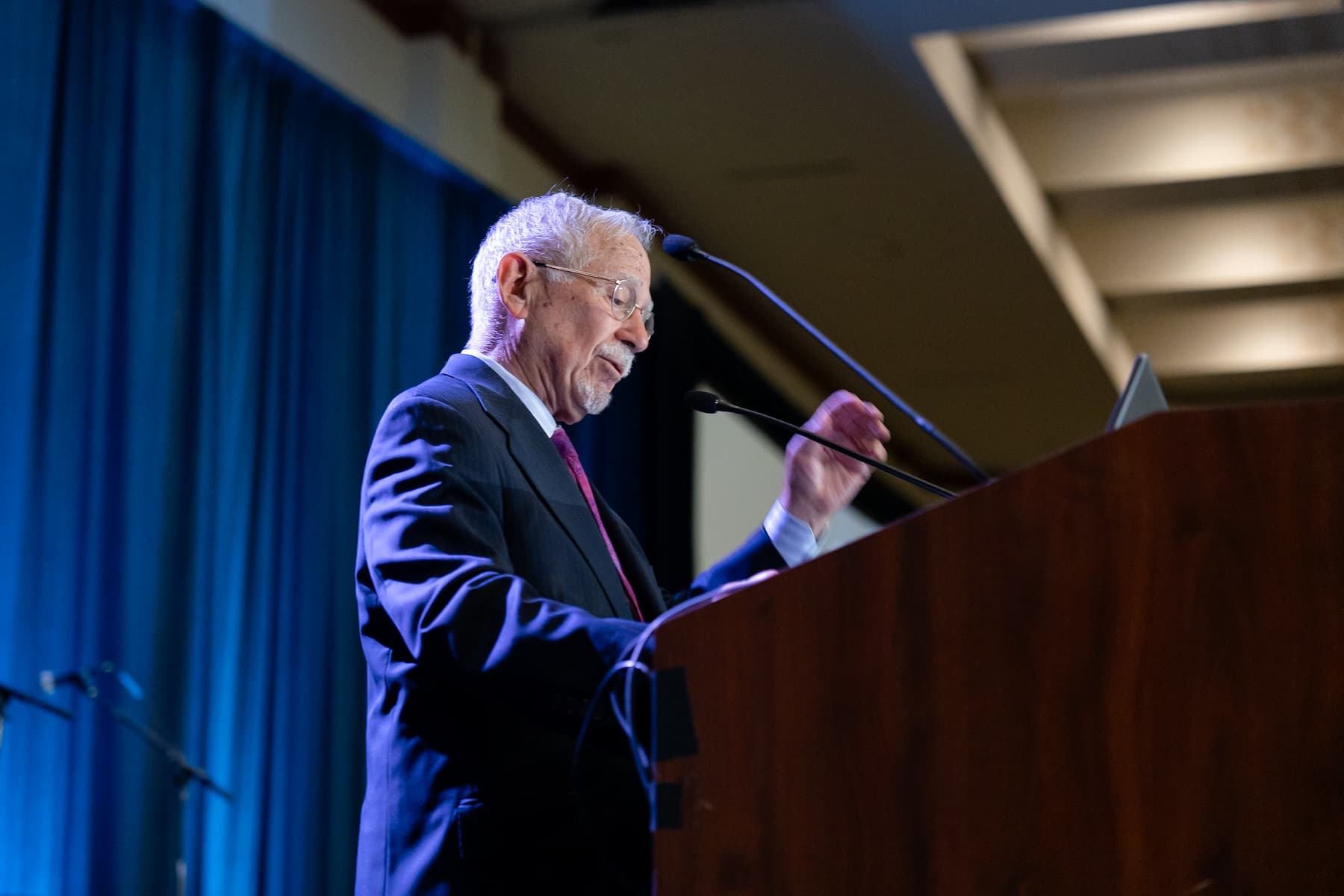 Person speaking at a podium during PAS 2025 conference, wearing a dark suit and red tie, with two microphones and a laptop visible; blue curtain backdrop and ceiling lights in the background.