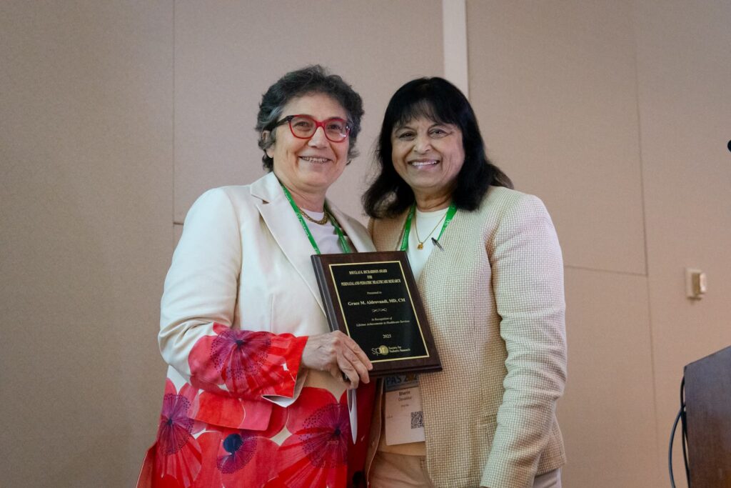 Two people standing indoors holding a plaque together, both wearing light-colored jackets and conference badges.