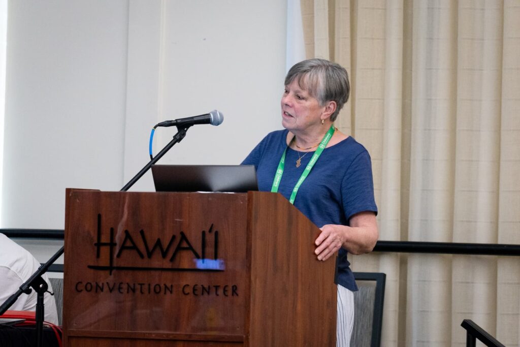 A person standing at a wooden podium labeled “Hawai‘i Convention Center,” delivering a presentation. The podium has a microphone and a laptop, and the individual is wearing a green lanyard. Beige curtains are visible in the background, suggesting an indoor conference setting.