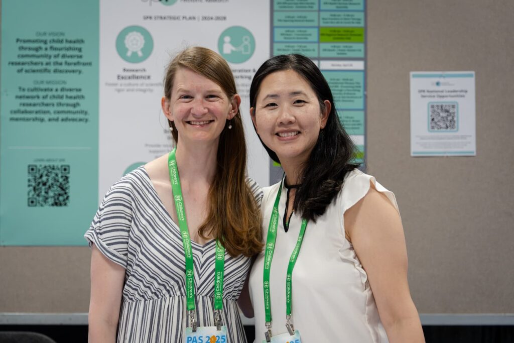 Two individuals standing in front of a poster display at a conference. Both are wearing green lanyards with badges, and the background includes a large poster featuring text, diagrams, and QR codes related to research or strategic plans.
