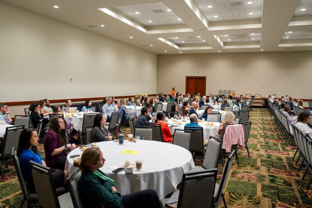 A large conference room with round tables and chairs arranged for a meeting or seminar. Several people are seated, facing the front of the room, with notebooks, drinks, and papers on the tables. The room has beige walls, patterned carpet, and a high ceiling with recessed lighting.