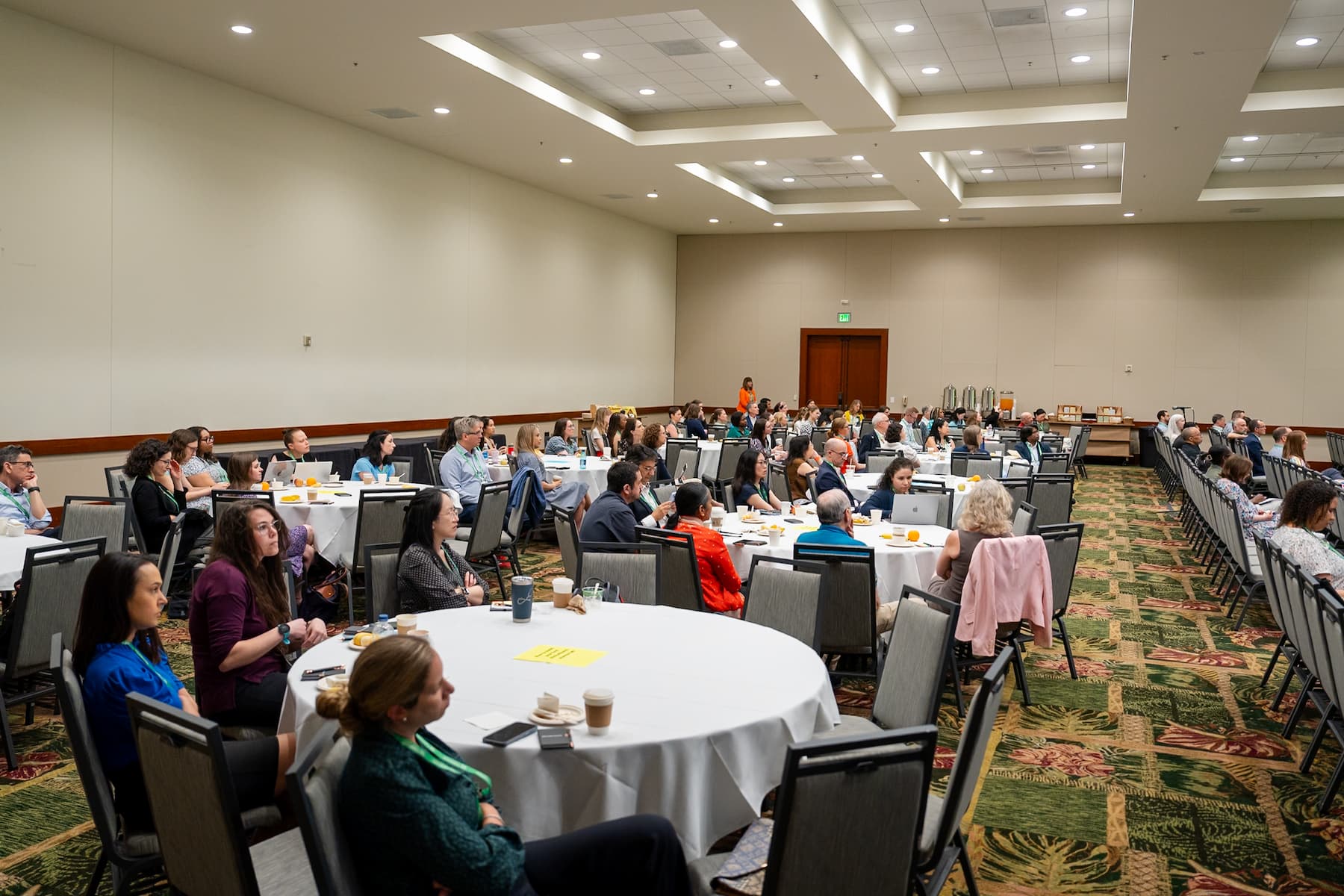 A large conference room with round tables and chairs arranged for a meeting or seminar. Several people are seated, facing the front of the room, with notebooks, drinks, and papers on the tables. The room has beige walls, patterned carpet, and a high ceiling with recessed lighting.