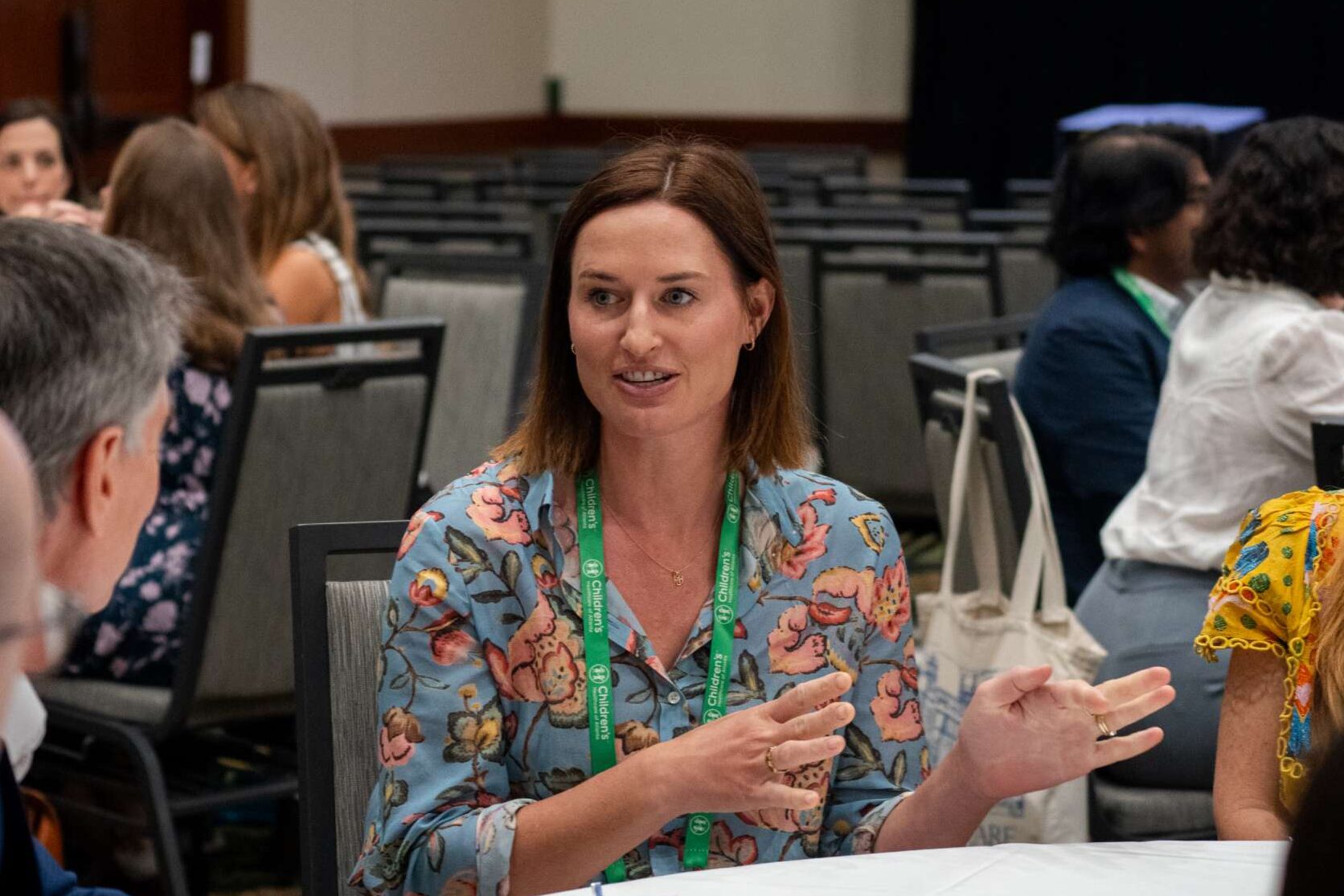 A person is seated at a table in a conference setting, engaged in conversation with others. The individual is wearing a floral-patterned top and a green lanyard with visible text. Several chairs and attendees are in the background, suggesting a breakout or networking session.