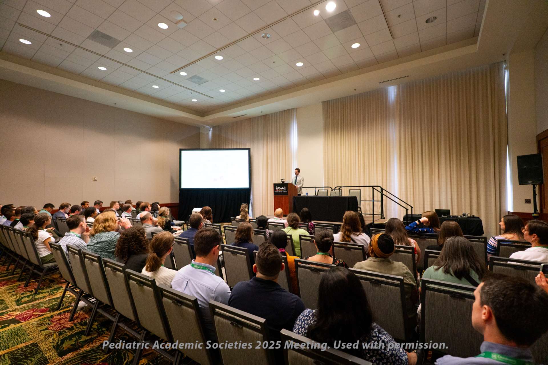 Audience seated in a conference room listening to a speaker at a podium, with a presentation screen displayed at the front and rows of chairs arranged facing the stage.
