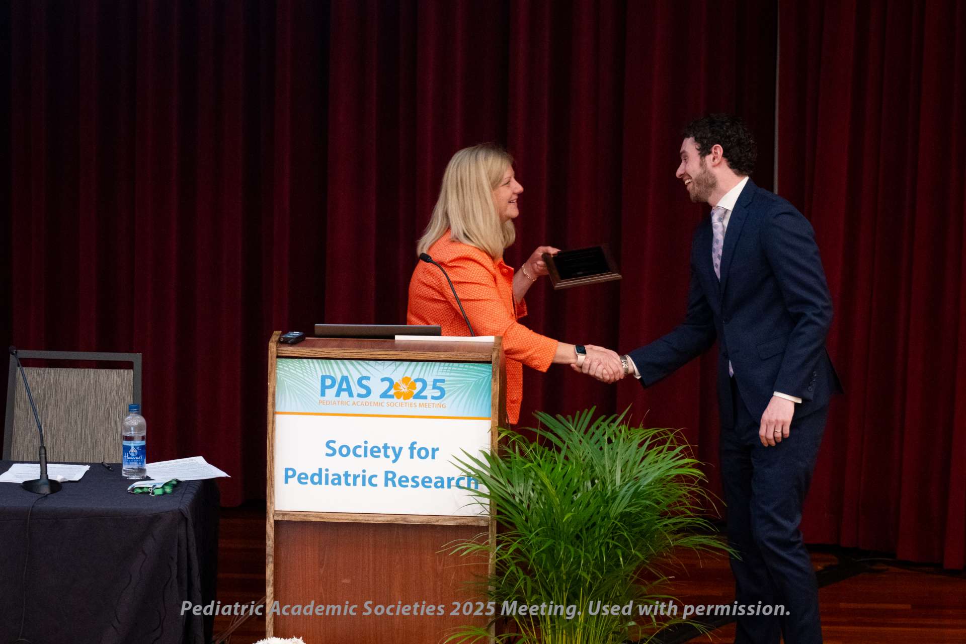 Two individuals shaking hands near a podium with a sign reading “PAS 2025 Society for Pediatric Research,” in front of a red curtain. One person is holding a plaque.