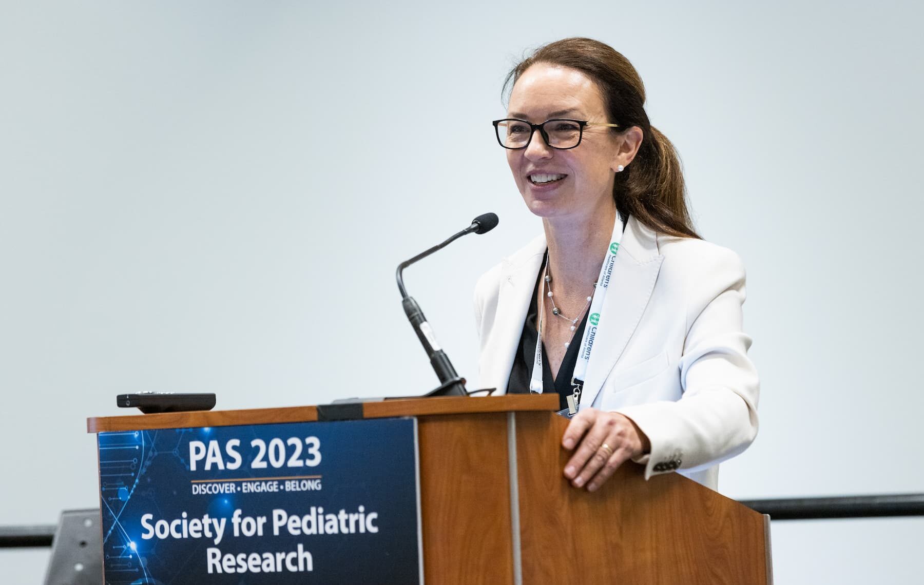 Person speaking at a podium during PAS 2023 conference, wearing a white blazer and green lanyard, with a microphone and a sign reading “PAS 2023 Society for Pediatric Research” visible on the podium.