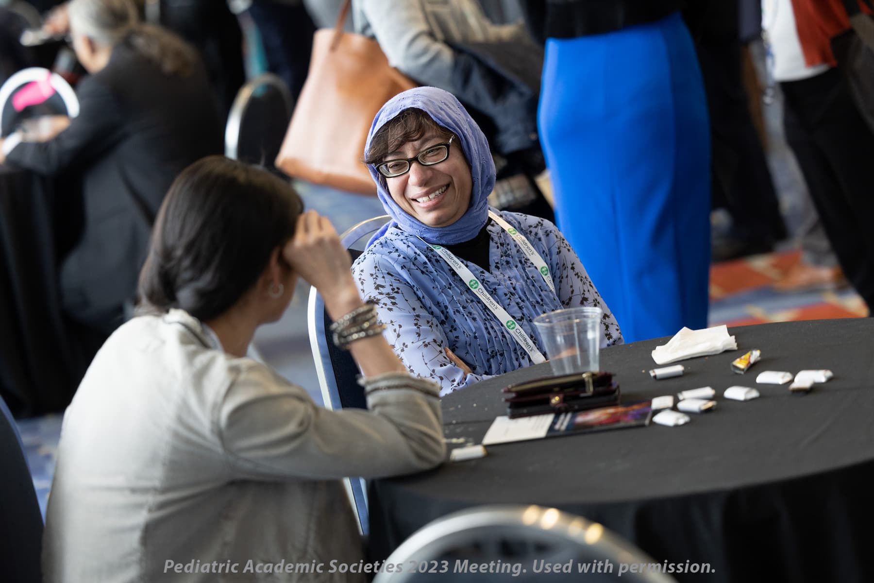 Two people seated at a round table during a conference or meeting, engaged in conversation. The table has a clear plastic cup, a smartphone, and several small items scattered on it. Other attendees and tables are visible in the background, suggesting a busy networking or discussion area.