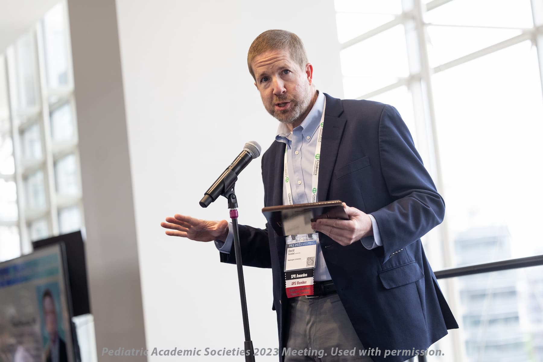 Person speaking into a microphone while holding a tablet at a conference, with large windows in the background.