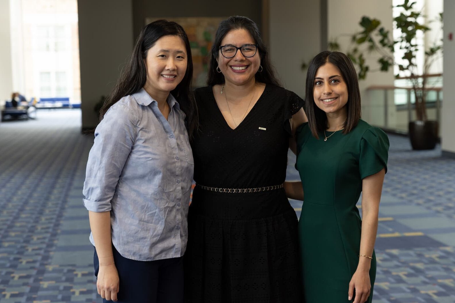 Three people standing together in a spacious indoor area with patterned carpet and large windows in the background. They are dressed in semi-formal attire, with one wearing a light button-up shirt, another in a black dress, and the third in a green dress. The setting appears to be a conference or meeting venue.