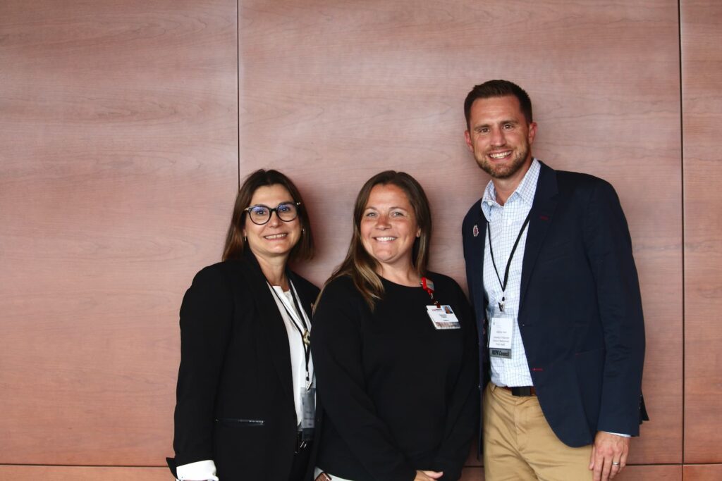 Three people standing together in front of a wooden wall, wearing business or conference attire with visible name badges and lanyards.
