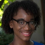 A person wearing glasses smiles in an outdoor setting with greenery in the background.