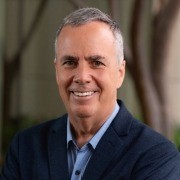 A person with short gray hair, wearing a dark blue blazer and light blue shirt, stands in front of a blurred outdoor background.