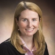 Person with long hair in a black jacket and pink shirt, smiling against a brown gradient background.