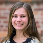 Person with long hair in a patterned sweater, smiling in front of a blurred brick background.