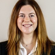 Person with long hair, wearing a white shirt and dark blazer, facing the camera against a plain light background.