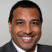 A person in a suit jacket and white shirt smiles against a plain light background.