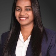 Person with long straight hair in a dark blazer and white shirt, standing against a plain dark background.