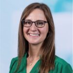 Person with long hair, wearing glasses and a green shirt, smiling at the camera against a light blue and white gradient background.
