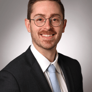 Person wearing glasses, a dark suit, white shirt, and light blue tie, standing against a plain gray background.