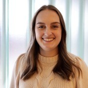 Person with long hair in a light sweater, smiling in front of a light blue curtain.