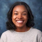 A person with dark hair, wearing a light sweater, smiles against a blurred blue-gray background.