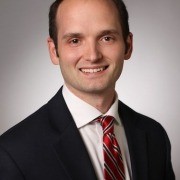 Person in a dark suit, white shirt, and red striped tie, standing against a plain light background.
