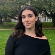 Person with dark wavy hair in a black off-the-shoulder top, standing outdoors with trees and buildings in the background.