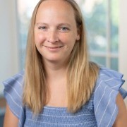Alt text: Person with long hair in a blue top with ruffled sleeves, standing indoors with blurred windows and greenery in the background.