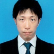 Person in a formal suit and tie with short dark hair, facing the camera against a plain blue background.
