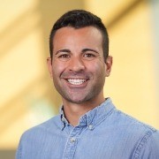 A person with short hair, wearing a light blue shirt, poses indoors with a blurred background.