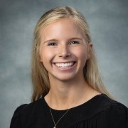 Person with long light hair in a dark top, smiling at the camera against a neutral gray background.