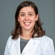 Person with shoulder-length hair in a white lab coat, smiling against a plain gray background.