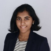 Person with short dark hair in a dark blazer and striped shirt, standing against a plain light background.