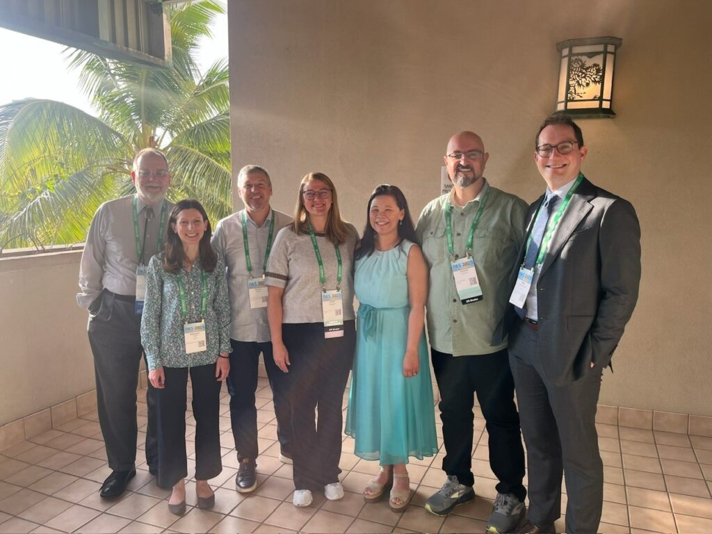 Group of seven people standing together on a tiled balcony at a conference, wearing event badges and green lanyards, with palm trees visible in the background and a wall lantern on the right.