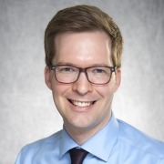 Person with short light brown hair wearing a light blue dress shirt and a dark tie, posed against a plain gray background.