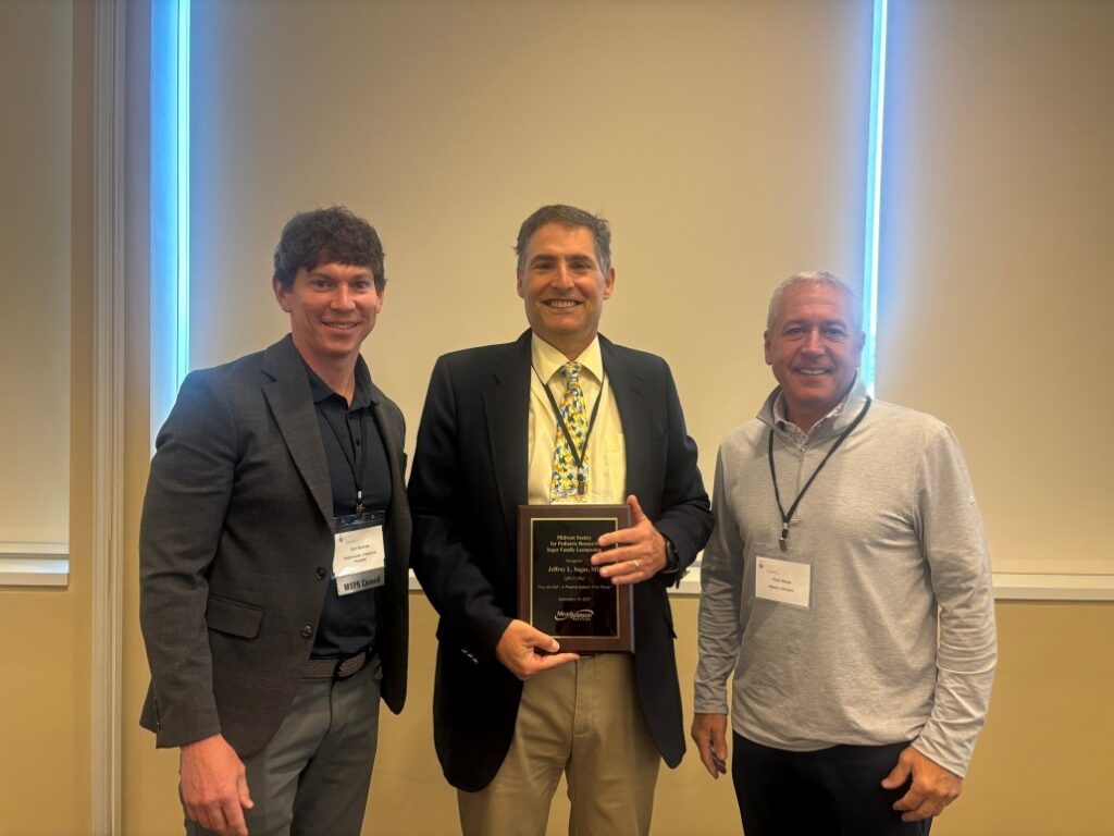 Four individuals standing indoors at a conference event. All are wearing business attire with name badges. One person in the center is holding a rectangular award plaque. Background includes beige walls and a projection screen