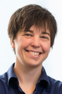 Person with short, tousled brown hair wearing a dark blue collared shirt, posed against a light background.