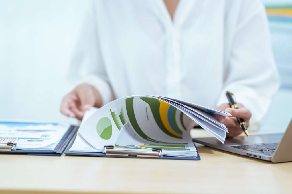 Person sitting at a desk reviewing a report with colorful charts and graphs on a clipboard, while holding a pen near an open laptop. Another clipboard with documents is placed on the desk.