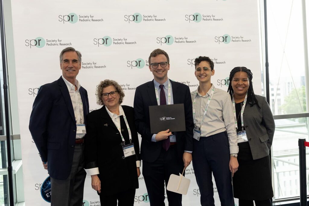 Five individuals in formal attire standing in front of a "Society for Pediatric Research" backdrop. One person in the center holds a black folder with the same logo.