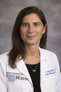 Person wearing a white medical coat with Emory Children’s and Children’s Healthcare of Atlanta logos, and a necklace, posed against a gray background.