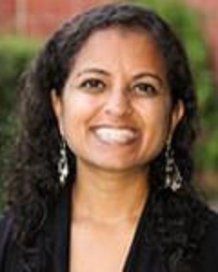 Person wearing a black top with long curly hair, standing outdoors with greenery and a brick wall in the background.