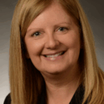 Person with straight shoulder-length blonde hair wearing a black top and a small round pendant necklace, smiling at the camera against a dark gray gradient background.