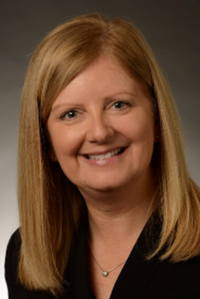 Person with straight shoulder-length blonde hair wearing a black top and a small round pendant necklace, smiling at the camera against a dark gray gradient background.